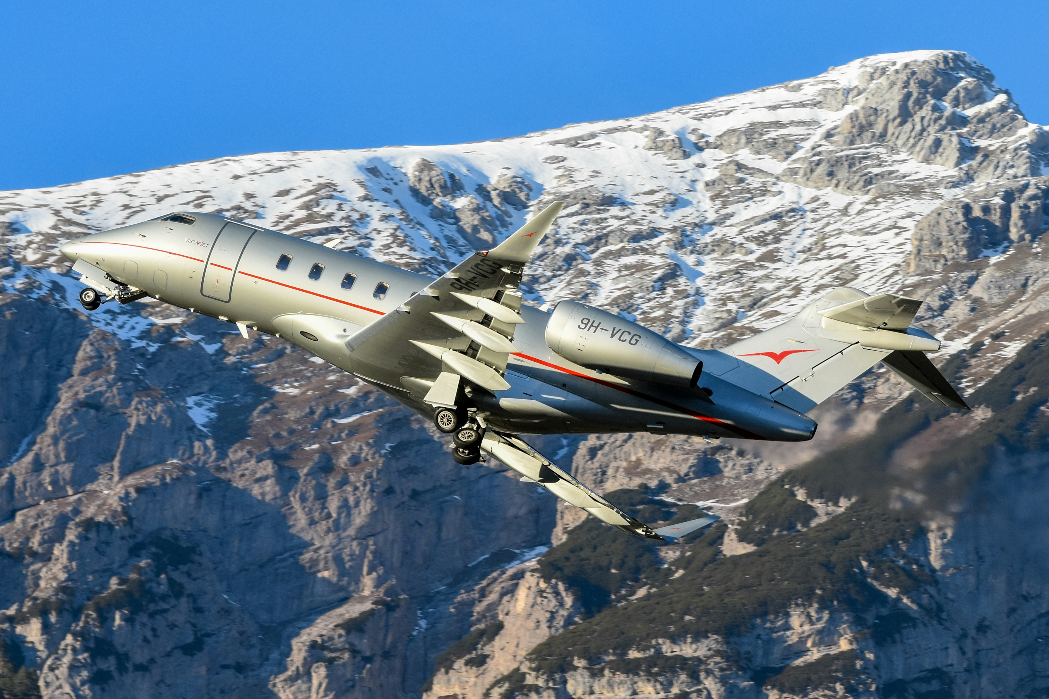 Silver private jet taking off against snow-dusted mountains.