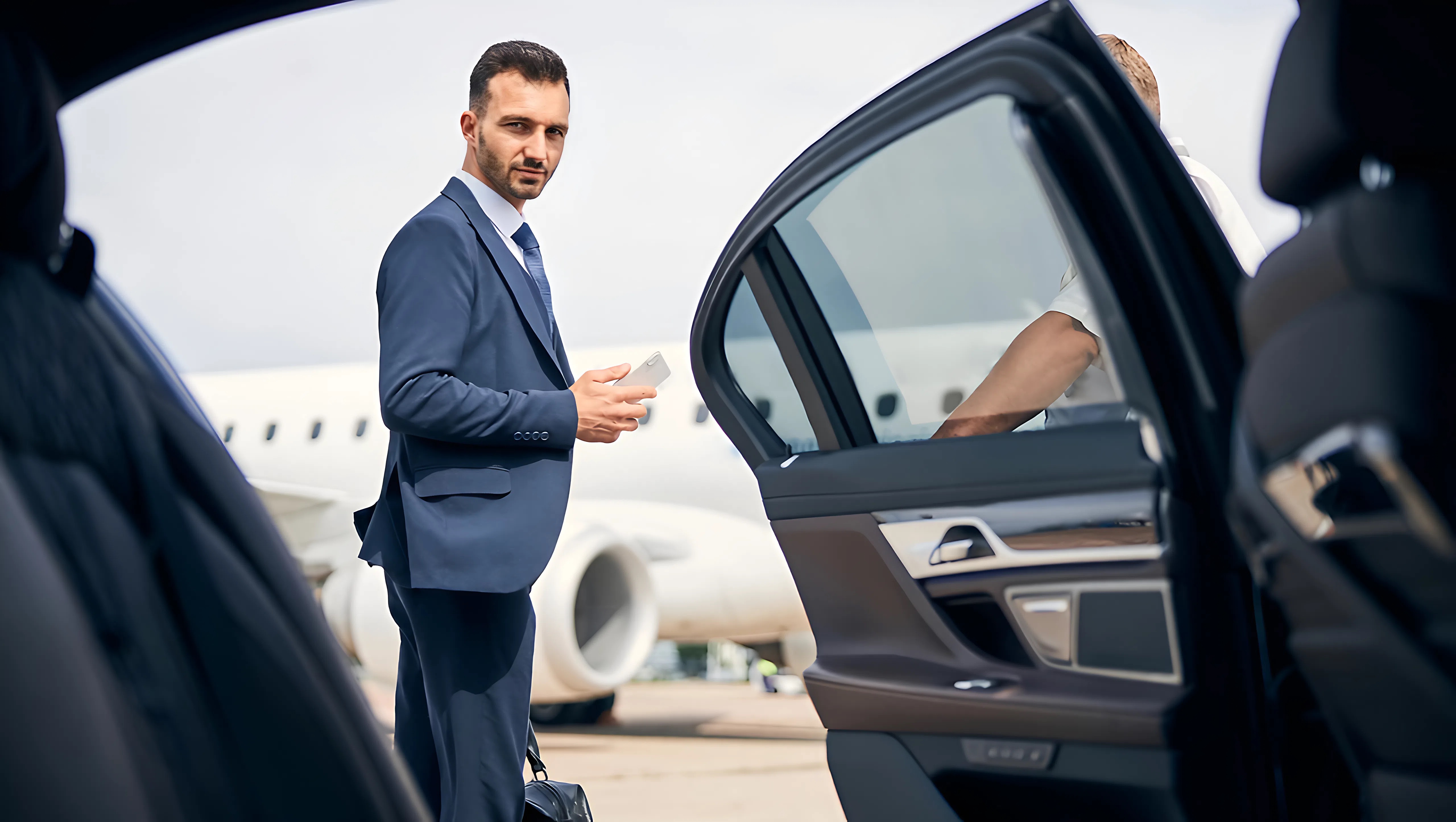 Businessman in a suit holding a phone by an open car door with private jet.